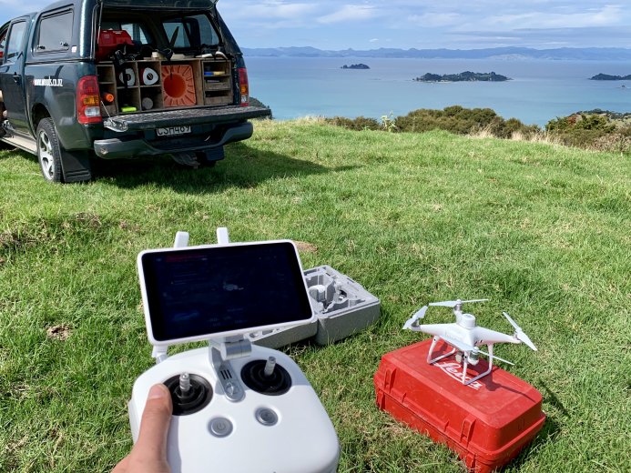 Small drone with remote control in hand, truck in foreground with harbour behind